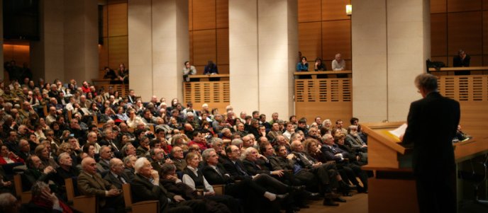 Professeur au pupitre donnant un cours devant un public dans un amphithéâtre.