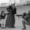 Femme âgée tenant deux fillettes par la main, faisant face à un soldat accroupi pointant une arme