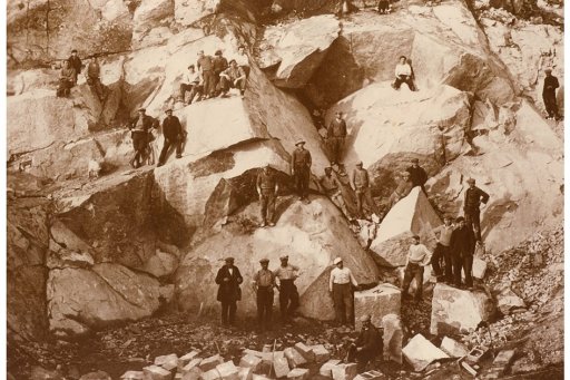 Photographie ancienne en sépia de travailleurs dans une carrière de pierre, dispersés sur des blocs rocheux avec des outils et des pierres taillées au sol.