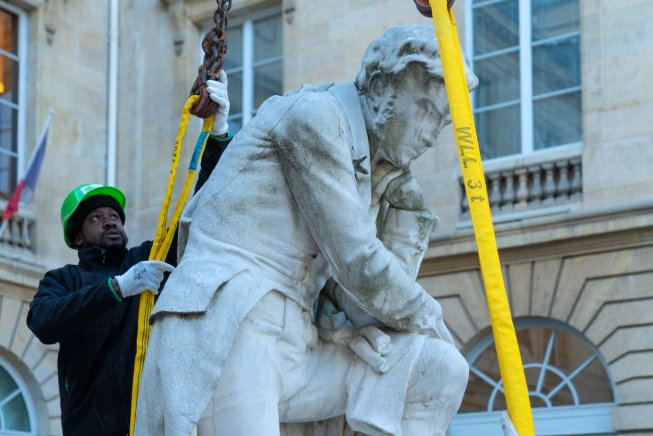 Ouvrier guidant une statue suspendue par des sangles jaunes devant un bâtiment