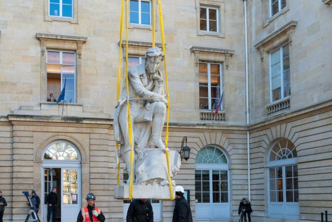 Statue suspendue par des sangles devant un bâtiment, entourée d'ouvriers et de spectateurs