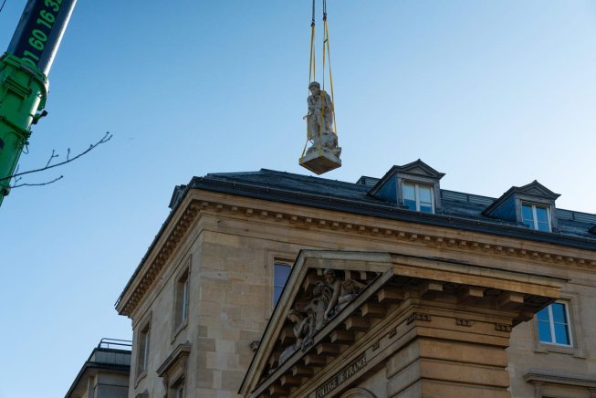 Statue suspendue par une grue au-dessus de l'entrée d'un bâtiment avec fronton sculpté