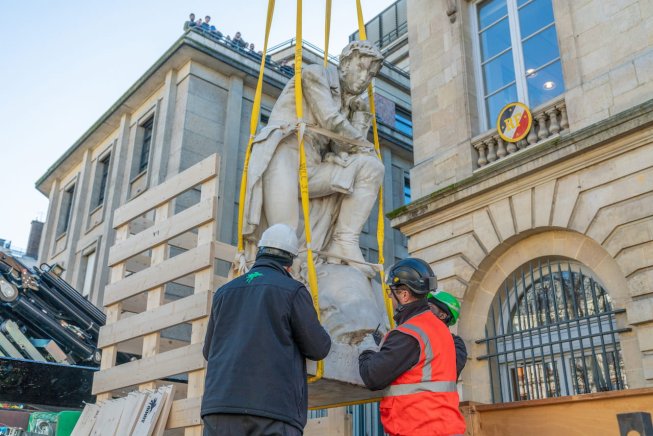 Statue suspendue descendue par des ouvriers avec des sangles devant un bâtiment