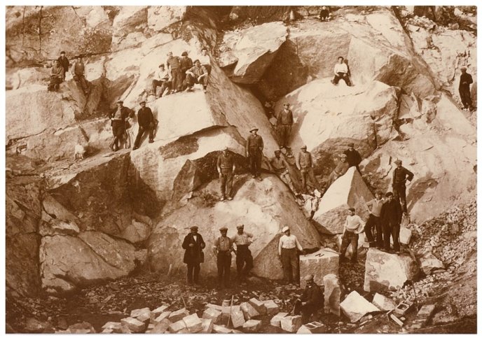 Early sepia photograph of workers in a stone quarry, scattered over boulders with tools and cut stones on the ground.