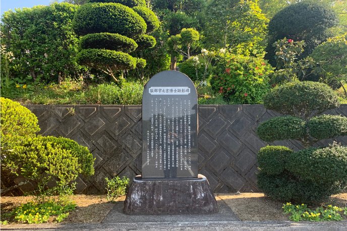 Stèle commémorative en pierre noire gravée de caractères japonais, érigée devant un mur de soutènement et entourée d’un jardin avec des arbustes et des plantes fleuries