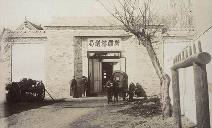 Photographie ancienne en noir et blanc montrant l'entrée d'un bâtiment traditionnel chinois en briques, surmontée d’une enseigne avec des caractères chinois signifiant "Bureau consultatif du Xinjiang" ; plusieurs personnes en vêtements d'époque se tiennent devant la porte ainsi qu’une charrette tirée par un cheval