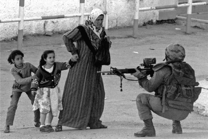 Femme âgée tenant deux fillettes par la main, faisant face à un soldat accroupi pointant une arme