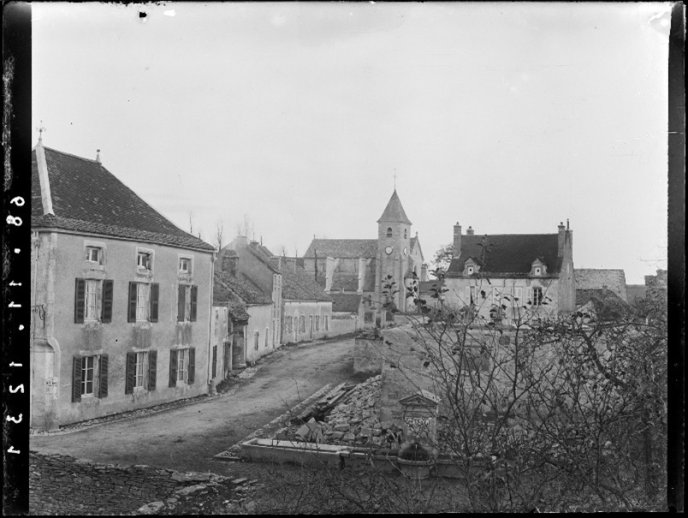 Vue ancienne d’un village avec église, maisons en pierre et fontaine centrale.