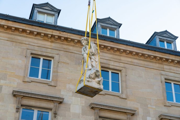 Statue en pierre suspendue par des sangles devant un bâtiment en pierre