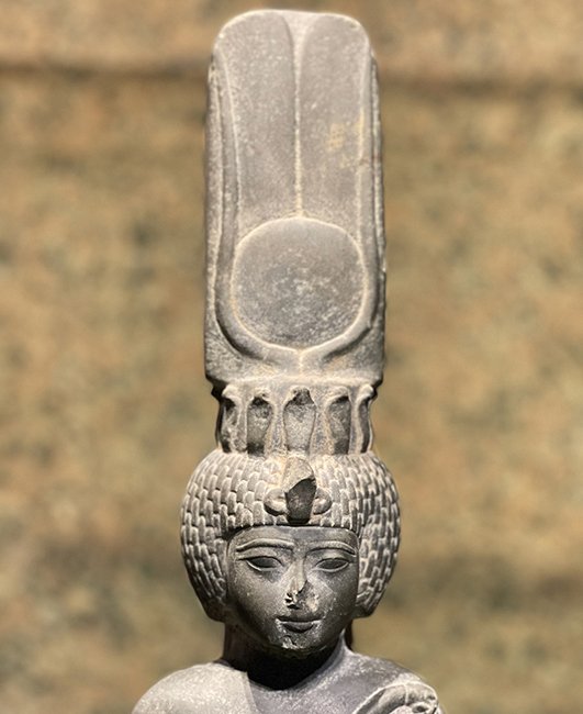 Display the full size picture: Statue représentant une divinité égyptienne, coiffée d’une haute couronne ornée, avec un visage sculpté finement détaillé