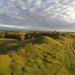 Paysage de collines herbeuses avec un chemin sinueux, entouré de champs et d’arbres sous un ciel nuageux